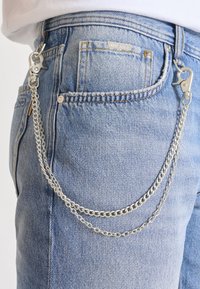 Accessorio in catena argentata attaccato a pantaloni in denim azzurro chiaro, con un moschettone e due catene collegate. Finitura testurizzata con un aspetto lucido.