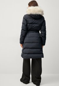 Navy blå pufferjakke med en faux fur hætte, der har en quiltet design og bælte i taljen. Glat tekstur med en knælang pasform.
