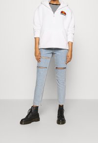 White fleece hoodie with front zipper and large pocket, layered over black and white striped shirt. Light blue ripped jeans and black boots.