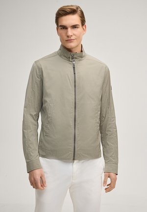 Junger Mann mit hellbraunen Haaren, der eine hellgraue Jacke mit Stehkragen und Reißverschluss sowie weiße Hosen trägt und vor einem einfarbigen Hintergrund steht.