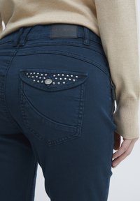 Donkerblauwe jeans met een slank gesneden ontwerp, voorzien van een rugzakje met sprankelende strasstenen en een gestructureerde tailleband met een logopatch.