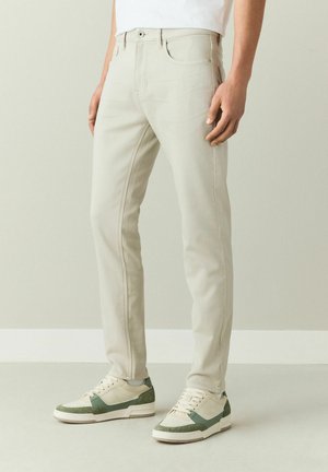 Mann trägt beige eng anliegende Hose und weiße Sneaker mit grünen Akzenten, steht vor einem hellen, schlichten Hintergrund.