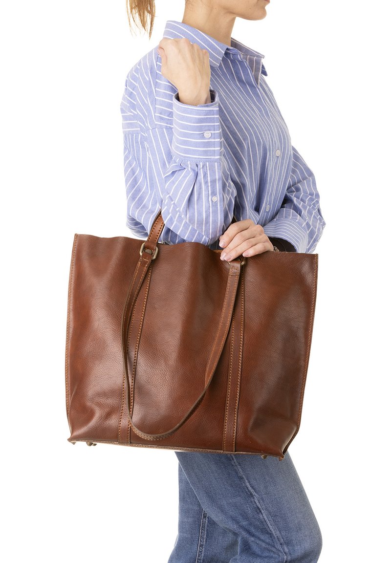 Sac en cuir brun avec deux longues anses, doté d'une texture lisse, d'une couleur profonde et de détails de couture subtils. Tenue par une personne.