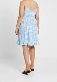 Frau in einem ärmellosen blau-weißen Batikkleid mit Rüschenumschlag und schwarzen Slipper-Sandalen, stehend vor einem schlichten Hintergrund.