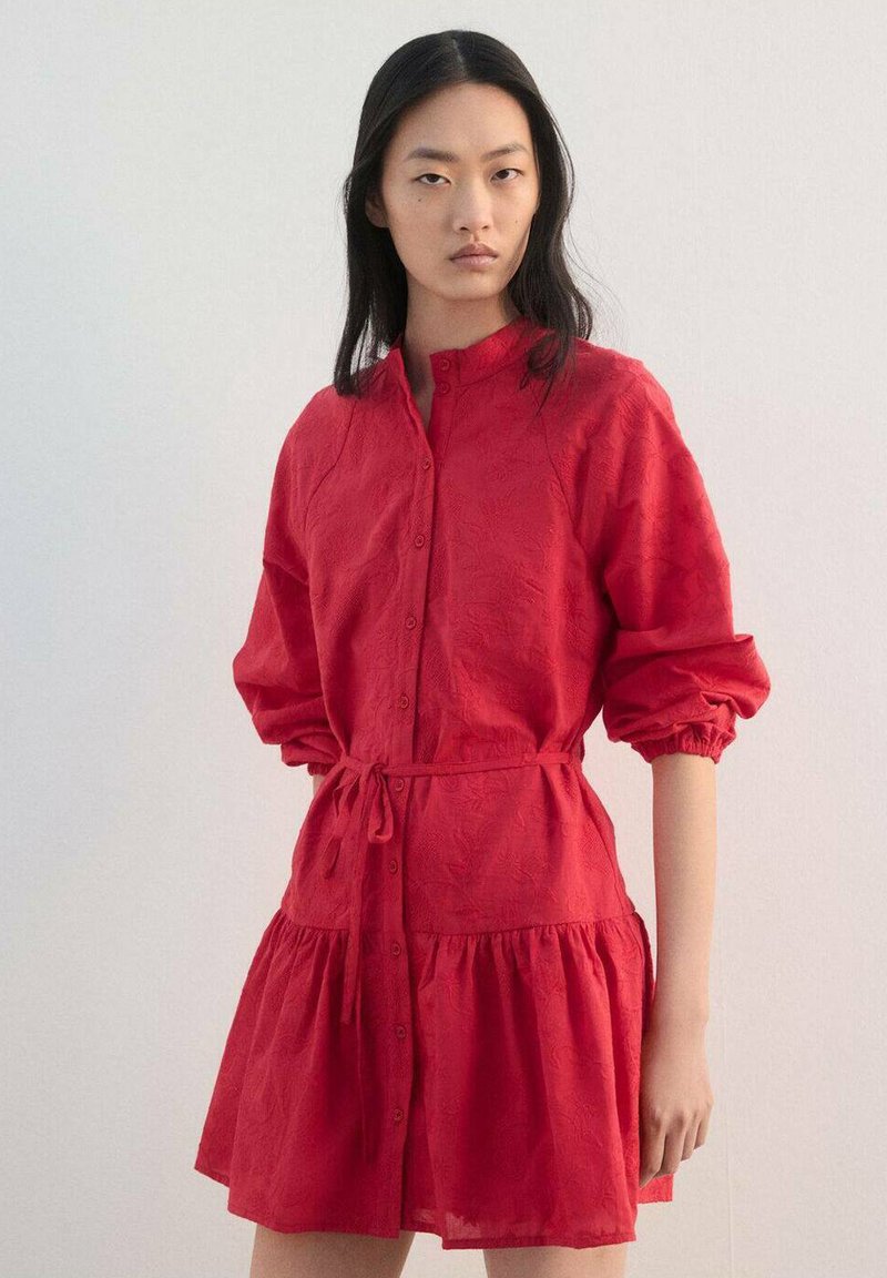 Jeune femme aux longs cheveux noirs portant une robe rouge à boutons avec manches longues et ceinture nouée à la taille, debout devant un fond uni.