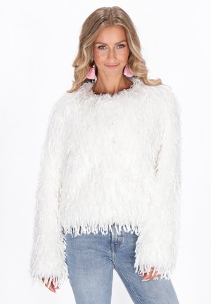 Un pull blanc duveteux avec une surface texturée et shaggy, bordé de franges. Il dispose de manches longues et d'une coupe décontractée, associé à un jean bleu.