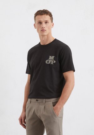 T-shirt nera in cotone con scollo rotondo, caratterizzata da un logo testurizzato in oro "M OP" sul lato sinistro del petto. Abbinata a pantaloni marrone chiaro.