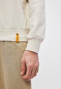 Κρέμα φούτερ με ριχτές μανσέτες, με κίτρινη ετικέτα με μαύρο κείμενο στο στρίφωμα. Χακί παντελόνι με ήπια υφή. Χέρι resting πάνω στο ύφασμα.