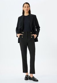 Zwarte blazer over een aansluitend zwart topje, gecombineerd met hoge zwarte broeken met gouden knoopdetails, en zwarte loafers met accenten.