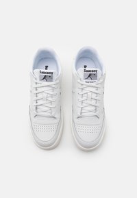 Zapatillas de cuero blanco con forro de malla, cordones planos y puntera perforada; presentan un logotipo negro en la lengüeta y una suela de goma.