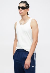 Top de alças branco de algodão com textura canelada, combinado com calças de fato de treino azul marinho com três riscas brancas e um logótipo na coxa. Óculos de sol pretos.