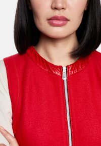 Rotes ärmelloses Kleidungsstück aus strukturiertem Stoff mit glänzendem roten Kragen. Verfügt über einen zentralen silbernen Reißverschluss und ein weiches, glattes Innenfutter.