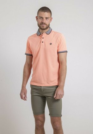 Homme portant un polo pêche avec col et poignets sombres, associé à un short vert olive retroussé, debout devant un fond uni.