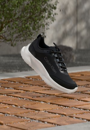 Zwart-witte Calvin Klein sneaker zweeft boven een oppervlak van houten tegels met groene bladeren en een grijze muur op de achtergrond.