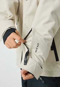 Beige waterproof jacket with adjustable cuffs, featuring black zippers and inner cuffs. Logo and STX 20.000 print on sleeve.