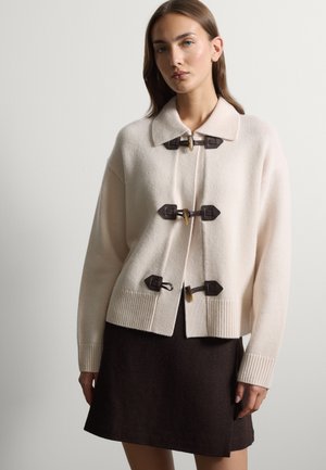 Donna che indossa un cardigan di maglia color crema con tre chiusure a bottoni scuri e una gonna marrone scuro su uno sfondo chiaro uniforme.