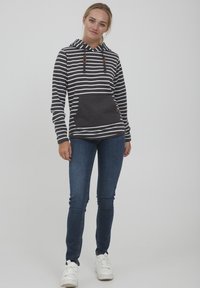 Stripet hettegenser i mørkegrå og hvit, med frontlomme og detaljer i treknapper. Kombinert med tettsittende blå jeans og hvite sko.