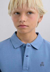 Lys blå polo skjorte lavet af struktureret stof med krave og tre knapper. Har et lille gråt kranium grafik på brystet.
