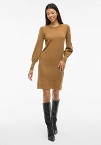 Long-sleeve, brown knitted dress with a round neck, fitted silhouette, and ribbed cuffs, paired with knee-high black boots.