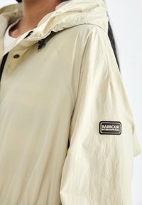 Világos bézs színű, kapucnis dzseki, texturált anyagból készült, bal ujja alatt logóval ellátott, valamint elöl cipzáros, patentgombos záródással.