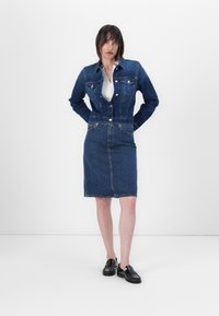 Jeansjakke med en tettsittende midje og et rett denimskjørt, begge i mørk blå. Har frontlommer og metallknapper. Enkel hvit topp under.
