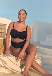 Costum de baie negru din două piese, cu detaliu strâns pe partea de sus, partea de jos cu talie înaltă și bretele subțiri. Model așezat pe o saltea împletită.