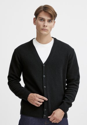 Cardigan noir en tricot avec un col en V, fermeture à six boutons sur le devant, poignets à côtes et tissu texturé, porté par-dessus une chemise blanche.