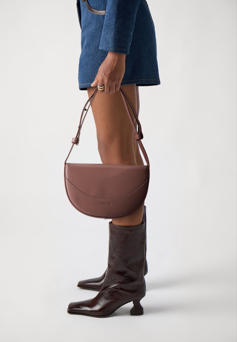Personne portant une jupe bleue, des bottes marron foncé jusqu'au milieu du mollet, tenant un sac à main marron en forme de demi-lune par la lanière sur un fond uni.