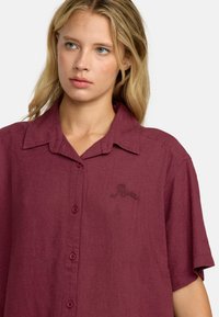 Camicia di lino color marsala con maniche corte, chiusura frontale con bottoni e testo ricamato in modo sottile sul lato sinistro del petto. Texture liscia e vestibilità rilassata.
