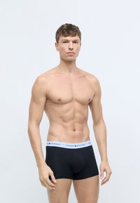 Sorte boxer briefs med en lys blå elastik, der har "TOMMY HILFIGER" logoet. Modellen har kort, brunt hår og en muskuløs krop.