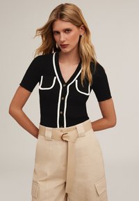 Czarny kardigan w prążki z białymi akcentami i guzikami, z dwiema kieszeniami na piersi. Połączony z beżowymi spodniami z wysokim stanem i paskiem.