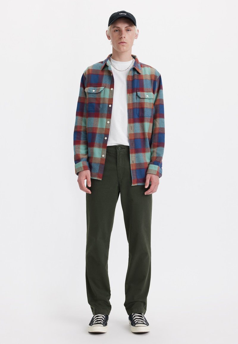 Camisa de franela con un patrón a cuadros multicolores en azul, rojo y verde, llevada sobre una camiseta blanca, combinada con pantalones verdes y zapatillas negras.