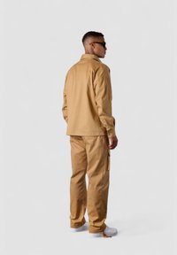 Khaki utility jacket and pants made of cotton, with a relaxed fit and cargo pockets. Paired with white sneakers. Back view against a neutral background.