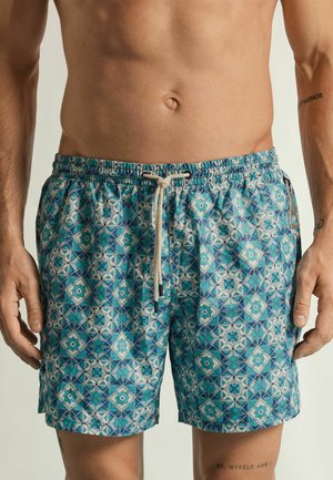 Buste masculin portant un short de bain bleu à motifs d'étoiles et de carreaux géométriques, taille élastique et cordon de serrage.