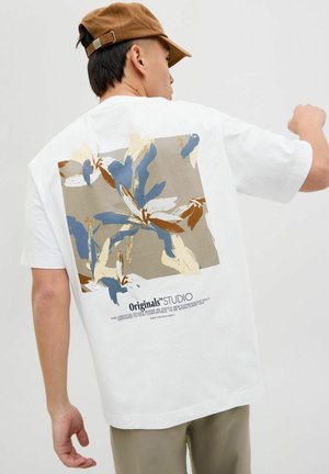 T-shirt blanc avec un grand imprimé floral en bleu, or et beige au dos, avec du texte en dessous du design. Manches courtes et coupe décontractée.