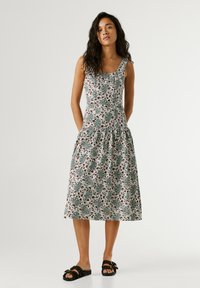 Mujer de pie vistiendo un vestido blanco sin mangas hasta la rodilla con un patrón floral verde y marrón y sandalias negras con hebillas.