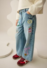 Jasnoniebieskie dżinsy z denimu o szerokich nogawkach, ozdobione różnokolorowymi haftowanymi naszywkami, w tym sercami i deserami. Noszone z czerwonymi trampkami.