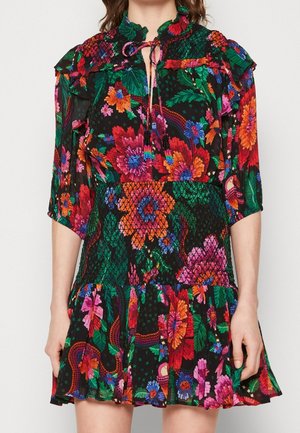 Kvinde iført en kort, sort blomsterkjole med levende pink, orange og grønne blomster, et smocket talje, flæsekanter og 3/4 ærmer.