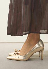 Zelta laku ādas augstpapēžu kurpes ar izvirzītiem pirkstiem, ar loku detaļu uz vampi un stiletto papēdi.