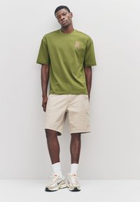 Olijfgroene T-shirt met korte mouwen en een logoprint; gecombineerd met beige korte broek. Model draagt witte sokken en beige sneakers met accenten.