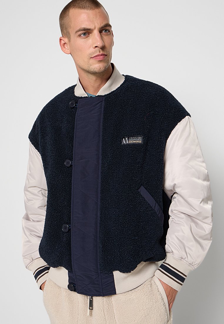 Giacca in pile blu navy con la parte inferiore trapuntata beige, dotata di bottoni a pressione, tasche laterali e una patch con il logo sul petto.