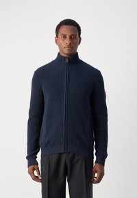 Maglione a coste navy con colletto alto e zip completa; presenta tasche laterali e un design aderente con polsini e orlo a coste.