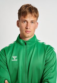 Veste de sport verte avec fermeture éclair sur le devant, fabriquée en tissu lisse. Présente un logo sur la poitrine et des accents blancs sur les épaules.