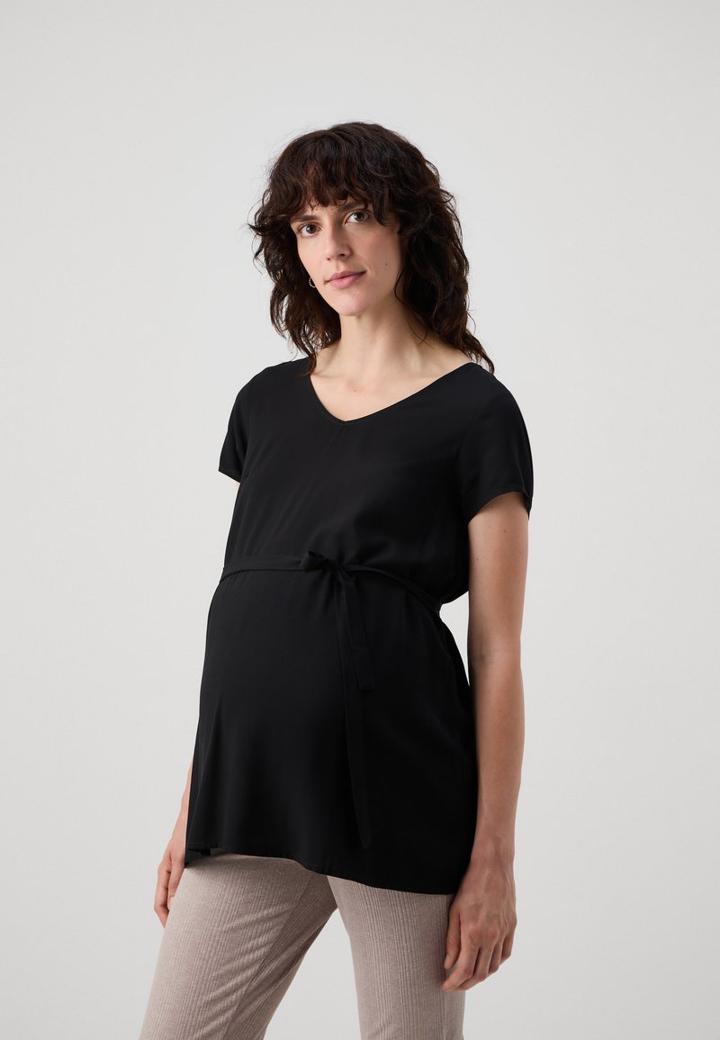 Top da maternità nero con scollatura a V e maniche corte, con dettaglio di vita annodata. Indossato con pantaloni beige aderenti e texturizzati.