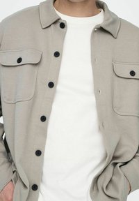 Light gray, textured button-up shirt with a collar, two chest pockets, and black buttons, layered over a white t-shirt.