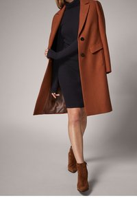 Manteau en laine rouille à double boutonnage porté sur une robe ajustée bleu marine, avec des manches longues et des boutons noirs. Bottines brunes à bout carré avec une finition texturée.