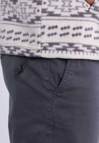 Giacca in pile color crema con motivi geometrici grigi, dotata di tasca con zip e dettaglio con cordino regolabile sull'orlo, abbinata a pantaloni scuri.