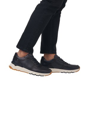 Black leather sneakers with textured accents, lace-up design, cushioned sole, and contrasting tan outsole. Worn with black denim pants.