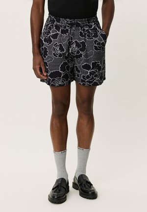 Szorty kąpielowe z nadrukiem kwiatowym w kolorach czarnym i szarym, wyposażone w elastyczny pas, boczne kieszenie oraz luźny krój. Zestawione z szarymi skarpetkami i czarnymi butami.