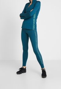 Top sportivo turchese con colletto alto e maniche lunghe abbinato a leggings turchesi, entrambi con dettagli laterali riflettenti, e sneaker nere.